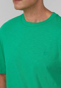 Zielona koszulka z krótkim rękawem, wykonana z miękkiego materiału, z okrągłym dekoltem i małym, subtelnym logo na piersiach. Gładka faktura, bez wzorów.