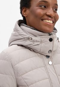 Lichtbeige pufferjack met een hoge kraag, horizontaal gestikt patroon, zilveren knopen en een grijze fleece voering zichtbaar bij de kraag.