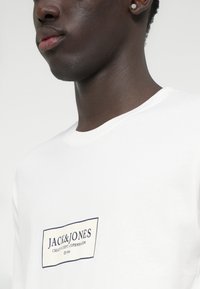 Detailný záber osoby v bielom tričku s logom "JACK & JONES Creative Dept. Copenhagen ©1990" na hrudi, na neutrálnom pozadí.