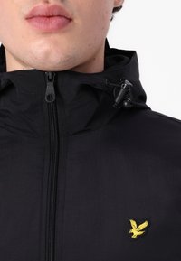 Zwarte lichtgewicht jack met een ritssluiting aan de voorkant, verstelbare capuchon en geel geborduurd logo. Heeft een gladde textuur en een minimalistisch design.