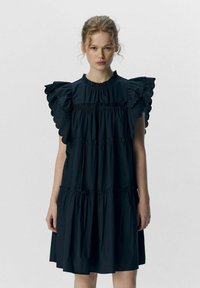 Schwarzes Baumwollenkleid mit Rüschennähten, hohem Ausschnitt und gestuften Design. Es hat eine weiche Textur und Falten im Taillen- und Saumbereich.