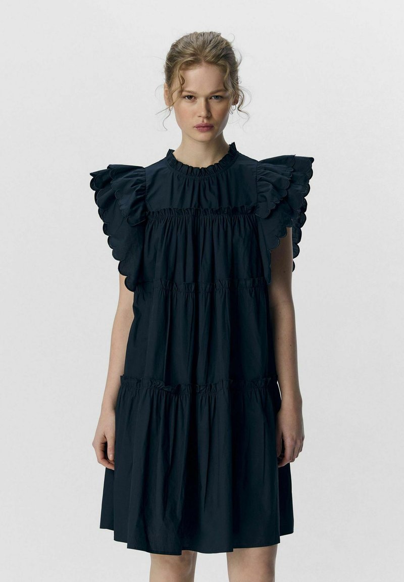 Schwarzes Baumwollenkleid mit Rüschennähten, hohem Ausschnitt und gestuften Design. Es hat eine weiche Textur und Falten im Taillen- und Saumbereich.