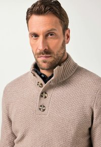 Muž s krátkými hnědými vlasy a vousy, oblečený v béžovém strukturovaném pleteném svetru s vysokým žebrovaným límcem a čtyřmi velkými knoflíky.