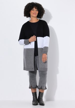Femme avec des cheveux bouclés portant un long cardigan noir, blanc et gris, un haut noir, un jean gris avec un ourlet effiloché, et des bottines noires à clous.