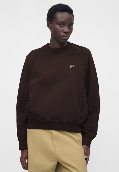 Άτομο που φοράει σκούρο καφέ φούτερ με μικρό κεντητό λογότυπο ζώου στο στήθος και ανοιχτό μπεζ παντελόνι, στέκεται μπροστά σε απλό φόντο.