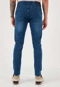 Sötétkék farmernadrág, slim fit fazonban, standard derékrésszel, két hátsó zsebbel és sima textúrával. Bézs cipővel viselve.