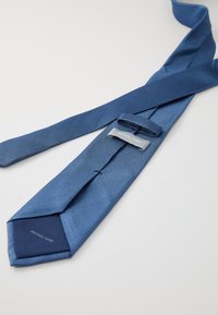 Blå slips med teksturert stoff, prydet med et geometrisk mønster. Avsmalnet form, med en etikett som viser "MICHAEL KORS" nær knuten.