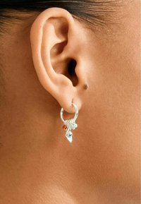 Nahaufnahme eines Ohrs mit einem silbernen Creolenohrring, der kleine herabhängende Charms in Blau, Bernstein und Silber zeigt.