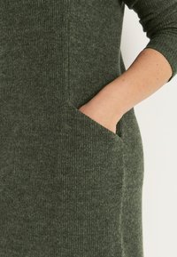 Stickad klänning i mörkgrön, med ribbat mönster och sidofickor. Fitted design med långa ärmar och mjukt tyg.