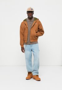 Brun hættejakke med grønt foer, grå polo-shirt, lyseblå jeans og lysebrune arbejdssko. Outfittet har afslappede byggeelementer.