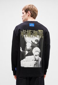 Μαύρο μπλουζάκι με μακριά μανίκια και γραφικό σχέδιο στο πίσω μέρος που απεικονίζει χαρακτήρες σε γκρι-κλίμακα, με κίτρινο κείμενο και μπλε ετικέτα στον ώμο.