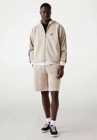 Beež värvi dressipluus lukuga ja küljetaskutega, sobitatud sobivate lühikestega. Omadeks mustad triibud ja logo. Mustad tossud valgete taldadega.