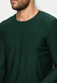 Ciemnozielona koszula z długim rękawem, uszyta z miękkiej tkaniny, z okrągłym dekoltem i dopasowanym krojem o gładkiej fakturze.