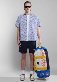 Korte mouwen, bloempatronen shirt in blauw en wit, gecombineerd met marineblauwe shorts. Model houdt een grote, multicolore sporttas met handvatten vast.