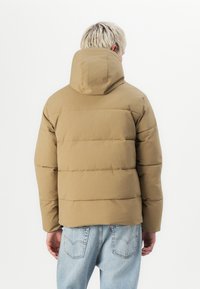Tan puffer jacket with a hood, featuring horizontal stitching and a matte finish. Shown from the back, paired with light blue jeans.