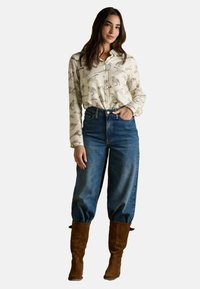 Woman wearing a cream button-up shirt with bird prints, high-waisted blue jeans, and brown suede boots, standing with one hand in pocket.