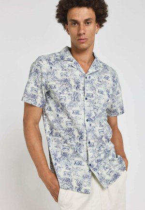 Homme aux cheveux bouclés portant une chemise à manches courtes avec un imprimé bleu d'animaux et de nature, mains dans les poches de pantalons blancs, fond neutre.