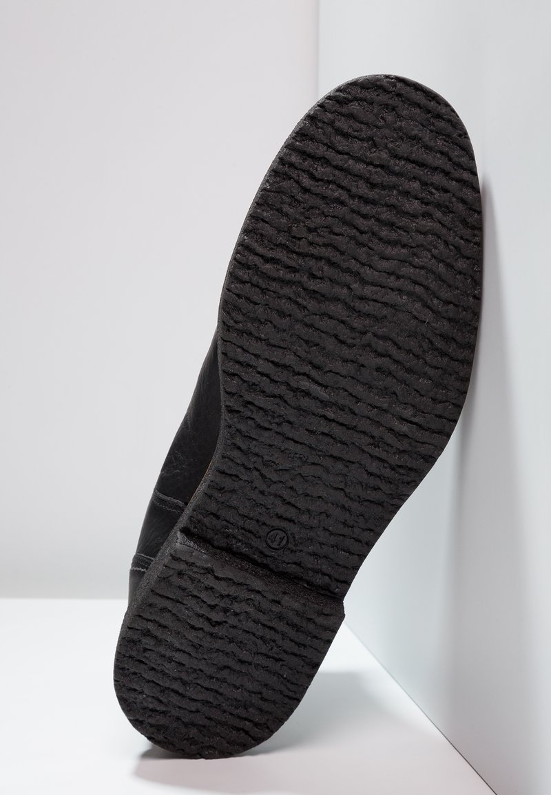 Suela de zapato negra con un patrón texturizado y estriado; sección superior parcial visible. El material parece ser de goma con un acabado suave.