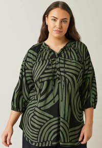 Blusa verde a fantasia con audaci strisce nere, scollo arricciato con fiocchi e maniche 3/4 con polsini elastici. Tessuto morbido.