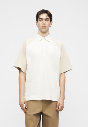 Νέος άνδρας με κοντά σκούρα μαλλιά, που φοράει διχρωμία μπεζ και άσπρο κοντομάνικο πόλο και ανοιχτό καφέ παντελόνι, στέκεται μπροστά σε λευκό φόντο.