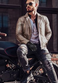 Giacca da motociclista in pelle tan, guanti neri, jeans scuri strappati, stivali neri. Seduto su una motocicletta nera, con occhiali da sole e una maglietta bianca.