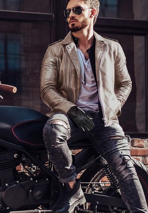 Chaqueta de moto de cuero marrón, guantes negros, jeans oscuros desgastados, botas negras. Sentado en una motocicleta negra, con gafas de sol y una camiseta blanca.