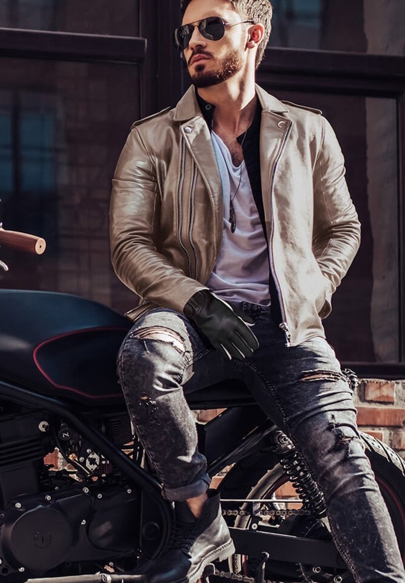 Giacca da motociclista in pelle tan, guanti neri, jeans scuri strappati, stivali neri. Seduto su una motocicletta nera, con occhiali da sole e una maglietta bianca.