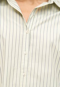 Κοντινό πλάνο ενός ατόμου που φοράει πουκάμισο εκρού με λεπτές μαύρες κάθετες ρίγες και τέσσερα λευκά κουμπιά, με τον γιακά ελαφρώς ανοιχτό.