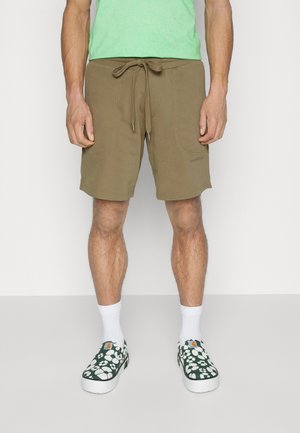 Homme portant un short vert olive à cordon, des chaussettes blanches et des chaussures vertes à enfiler avec un motif floral blanc, debout devant un fond uni.