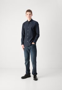 Marineblauwe overhemd met knopen en een logo, gecombineerd met donkere spijkerbroek en zwarte schoenen. Gladde stof, klassieke kraag en casual pasvorm.