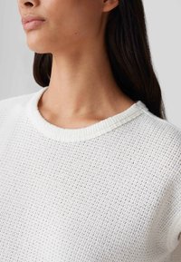 Witte, gestructureerde breiselsweater met een ronde hals en ribbeltrim, die een losse weefpatroon toont tegen een gladde huid.