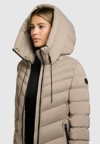Donna che indossa un giubbotto imbottito beige con un grande cappuccio, cerniera nera e lacci neri, che guarda a sinistra su uno sfondo neutro.