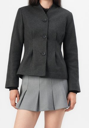 Kvinde iført en mørkegrå knaplukket blazer med skræddersyet pasform, kombineret med et lyst gråt plisseret skørt.