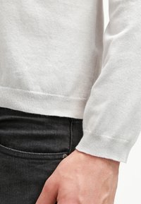 Jasnoszary sweter dzianinowy z prążkowanymi mankietami i dołem, o miękkiej fakturze; zestawiony z ciemnoszarymi spodniami z detalami bocznych kieszeni.