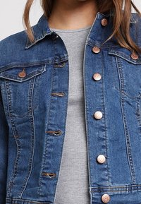 Džínová bunda, modrá, s knoflíkovým zapínáním, měděné knoflíky, dvě náprsní kapsy, mírně opotřebovaný vzhled, nošena přes šedý top.