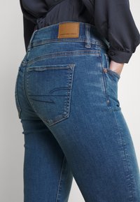 Denimjeans i medelblå färg med en slät yta. Har en läderlapp med texten "AMERICAN EAGLE" och sömmar på bakfickorna.