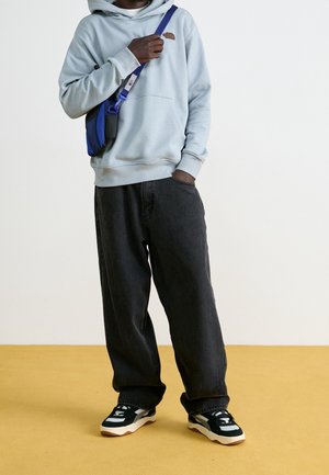 Person wearing a light gray hoodie, black wide-leg jeans, black and white sneakers, and a blue crossbody bag, standing on a yellow floor.