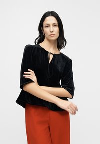 Black velvet blouse with a round neckline, short puff sleeves, and a drawstring detail at the front, paired with wide red trousers.