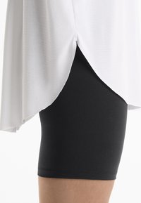 Haut blanc allongé avec un ourlet incurvé associé à un short noir ajusté. Le haut a une texture lisse; le short est extensible et moulant.