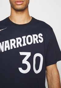 Homme portant un maillot bleu foncé des Golden State Warriors avec le numéro 30 et le logo Nike sur la poitrine, cadré au niveau du cou et du bras droit.