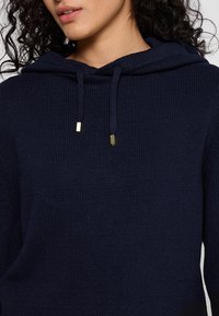 Marineblau gestrickter Hoodie mit strukturiertem Finish, ausgestattet mit einer Kordel mit goldenen Akzenten und einem lockeren Schnitt.
