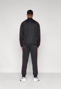 Μαύρη φόρμα από λείο ύφασμα με κόκκινες ρίγες στο πλάι, με ψηλό γιακά και ριπ μανσέτες, συνδυασμένη με άσπρα αθλητικά παπούτσια.