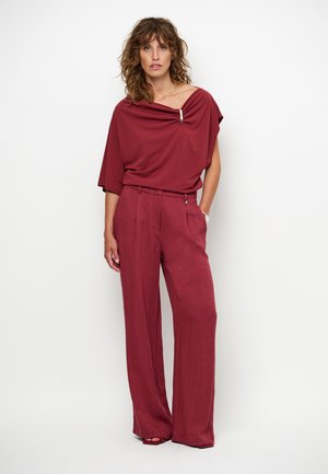 Blouse bordeaux avec décolleté drapé et accent métallique, associée à un pantalon large bordeaux taille haute. Tissu lisse, coupe décontractée.