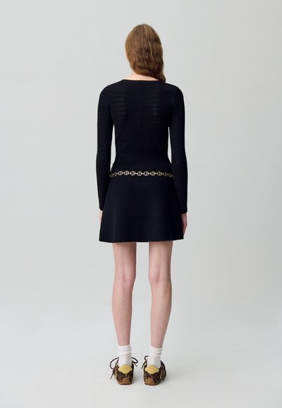Vestido negro de punto con mangas largas, que cuenta con una falda negra a juego y un cinturón decorativo de cadena dorada. Persona lleva zapatos con estampado.