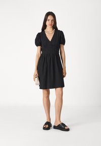 Μαύρο πλισέ φόρεμα με V λαιμόκοψη, κοντά φούσκωμα μανίκια και εφαρμοστή μέση. Συμπληρωμένο με μια πέρλα κολιέ και μαύρες πλατφόρμες.