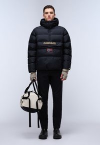 Giubbotto imbottito nero con cappuccio, caratterizzato da una toppa sul petto con logo e bandiera. Il modello tiene una borsa a tracolla beige e nera, indossando pantaloni scuri e stivali.