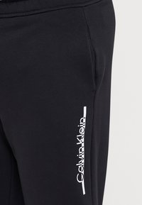 Schwarze Baumwoll-Jogginghosen mit einem weißen vertikalen Logo im Bereich der linken Tasche. Der Stoff hat eine weiche Textur und einen klassischen athletischen Schnitt.