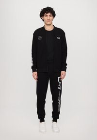 Black zip-up jacket with logo on chest, paired with black jogger pants featuring logo print. Both are made of a soft fabric. White sneakers.