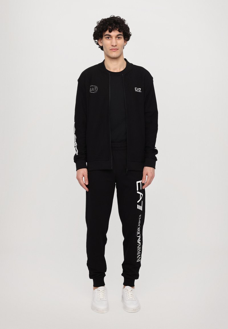 Chaqueta negra con cremallera y logo en el pecho, combinada con pantalones jogger negros con estampado de logo. Ambos están hechos de una tela suave. Zapatillas blancas.