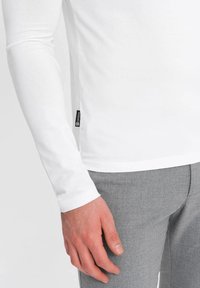 Witte blouse met lange mouwen en een zachte textuur, voorzien van een zwart label aan de zijkant, gecombineerd met lichtgrijze broek.